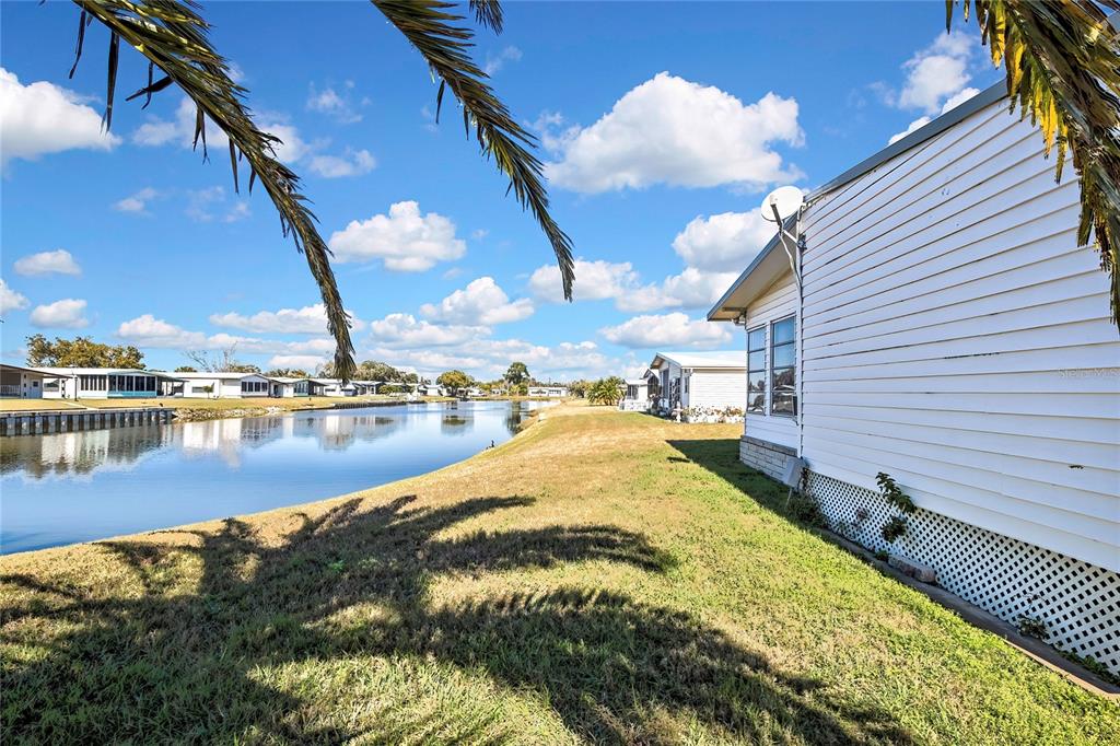 A white mobile home sits beside a canal with palm tree shadows on the grass — 3141 NANTUCKET DRIVE, HOLIDAY, FL