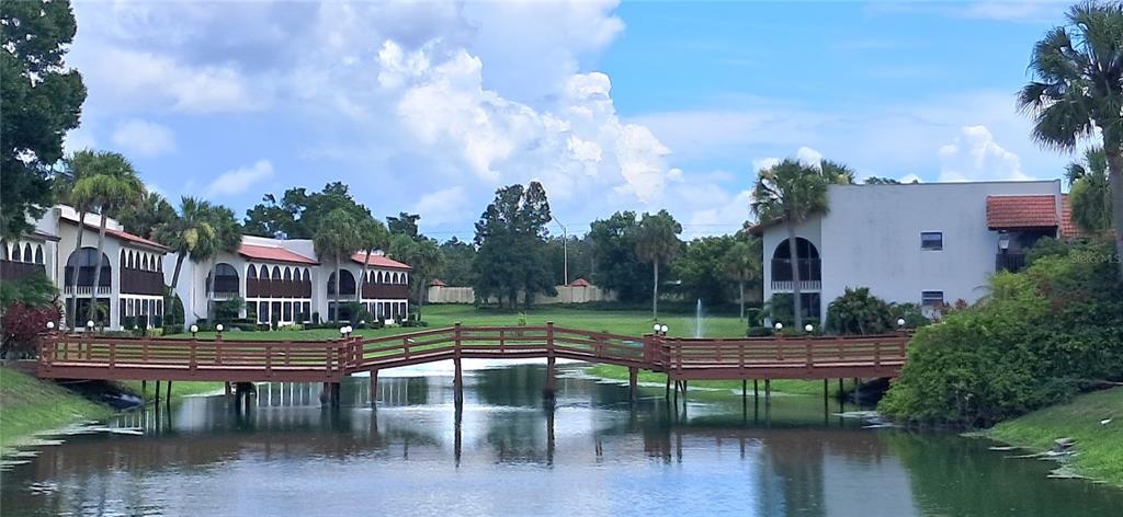 A wooden footbridge spans a pond in front of multi-story residential buildings with palm trees — 7201 COUNTRY CLUB DRIVE UNIT G21, LAKE WALES, FL