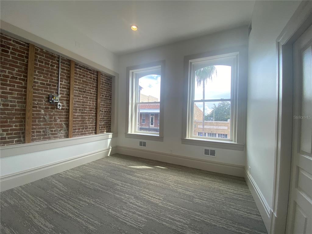An empty room features exposed brick walls, two large windows, and gray patterned carpeting — 1718 E 7TH AVENUE UNIT 201-C & E, TAMPA, FL