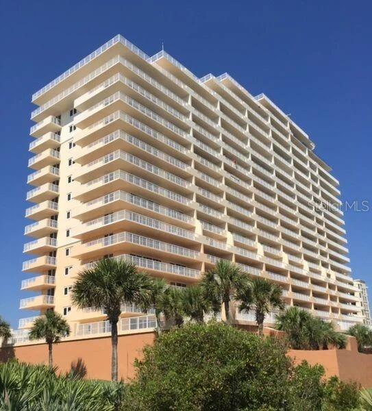 A tall, multi-story residential building with wrap-around balconies stands against a clear blue sky — 257 MINORCA BEACH WAY UNIT 1202, NEW SMYRNA BEACH, FL