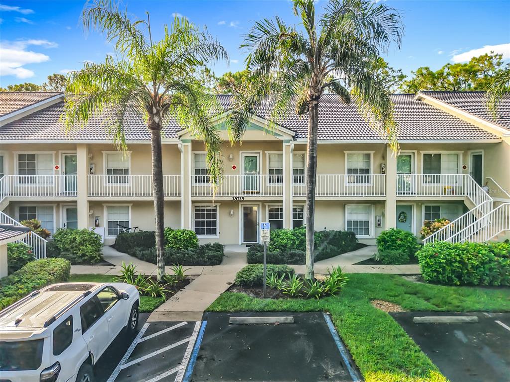 A two-story apartment building exterior features tan stucco walls, white railings, and manicured green landscaping — 25735 LAKE AMELIA WAY UNIT 103, BONITA SPRINGS, FL