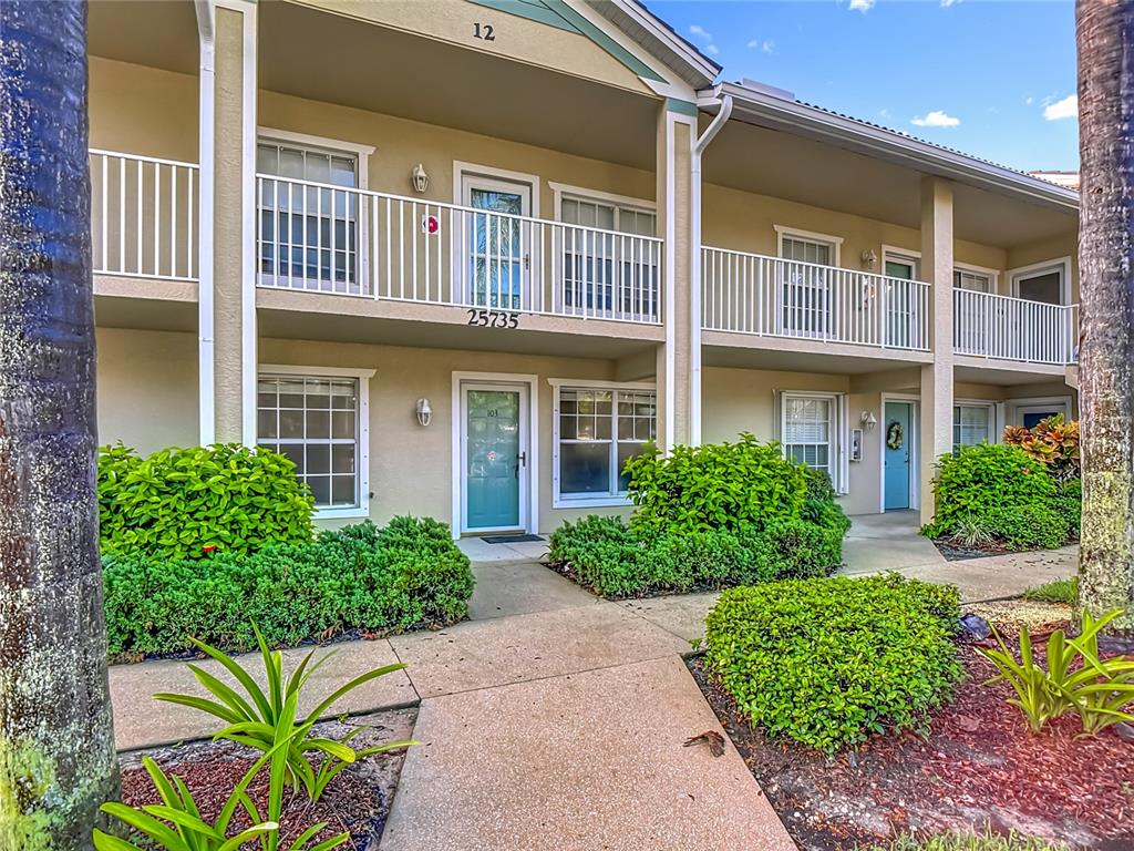 A ground-level building entrance features a teal front door, two windows, and surrounding landscaping — 25735 LAKE AMELIA WAY UNIT 103, BONITA SPRINGS, FL