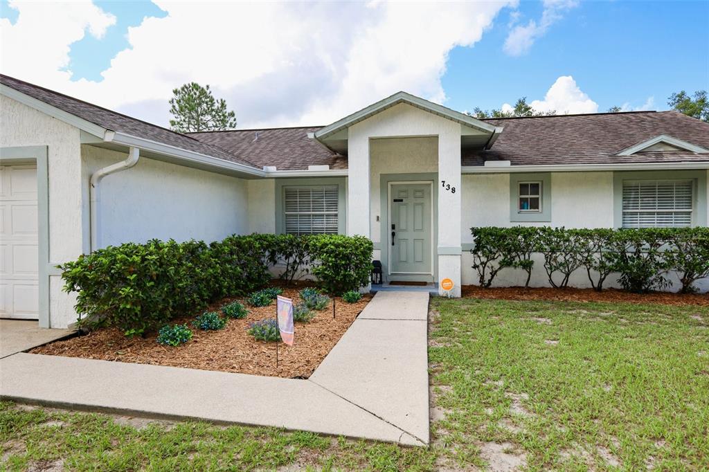 The exterior of a single-story house features white stucco walls, a brown roof, and landscaping — 738 E 11TH AVENUE, MOUNT DORA, FL