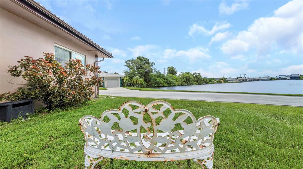 A white metal bench sits on a grassy lawn beside a house facing a lake — 5355 FERNDALE PLACE N, PINELLAS PARK, FL