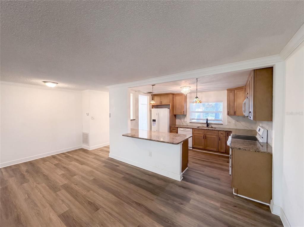 An open-concept kitchen with wood cabinets, granite countertops, and wood-look flooring overlooks a dining area — 5122 BAY BOULEVARD, PORT RICHEY, FL