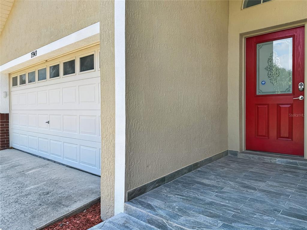 A red front door sits next to a white garage door on a tiled porch — 15943 HERON HILL STREET, CLERMONT, FL