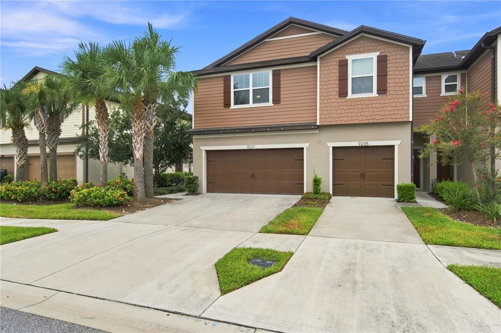 A two-story townhouse with brown siding, two garage doors, and palm trees in the front yard — 9237 HILLCROFT DRIVE, RIVERVIEW, FL