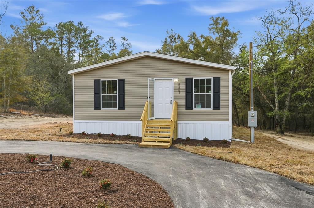 A tan manufactured home with black shutters and wooden stairs sits on a paved driveway — 25924 WHIPPERWILL STREET, BROOKSVILLE, FL