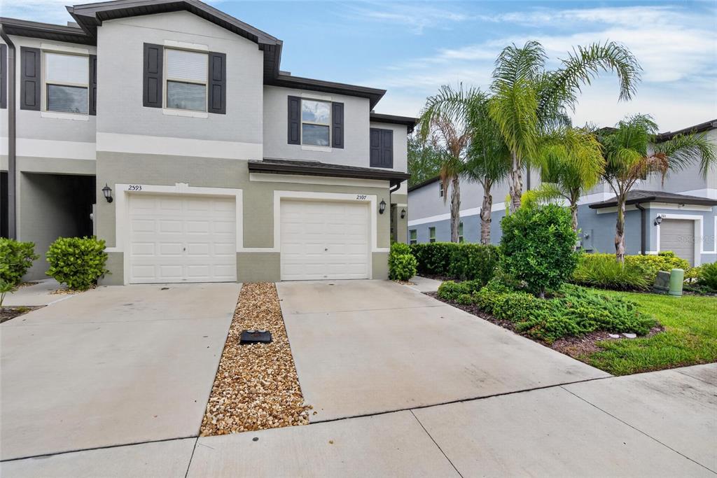 A two-story townhouse exterior features two garage doors, a gravel strip, and palm trees — 2597 CHARLAN, HOLIDAY, FL