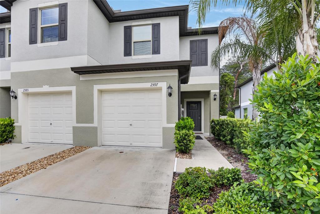A two-story townhouse exterior features a white garage door, a front entrance, and landscaping — 2597 CHARLAN, HOLIDAY, FL