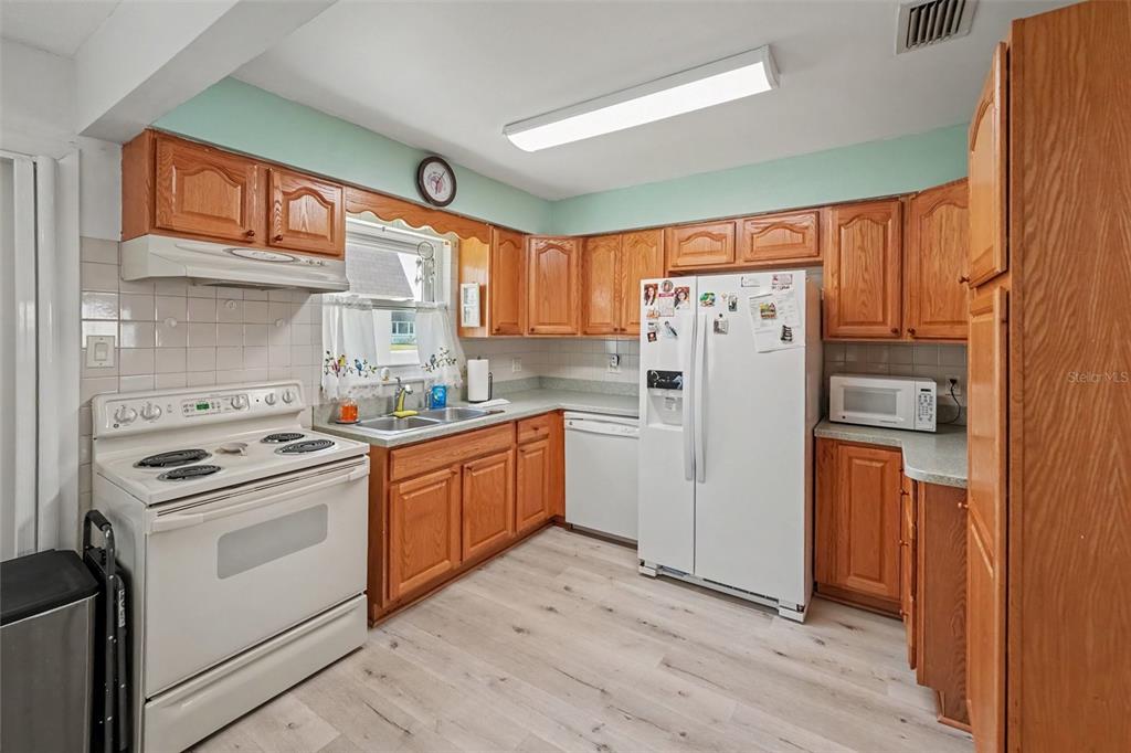 A kitchen with wood cabinets, white appliances, light-colored flooring, and a pale green wall accent — 6880 LAFAYETTE N UNIT 8, PINELLAS PARK, FL