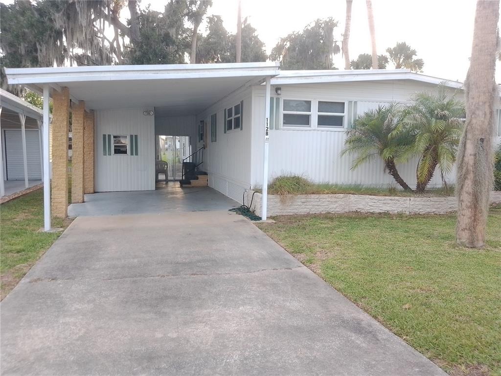 A single-story mobile home features a concrete driveway, a covered carport, and a small palm tree — 112 PALM DRIVE UNIT B, EUSTIS, FL