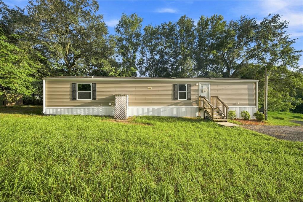 A tan mobile home with white skirting sits on a grassy lot with trees behind — 11945 SE 99TH COURT, BELLEVIEW, FL