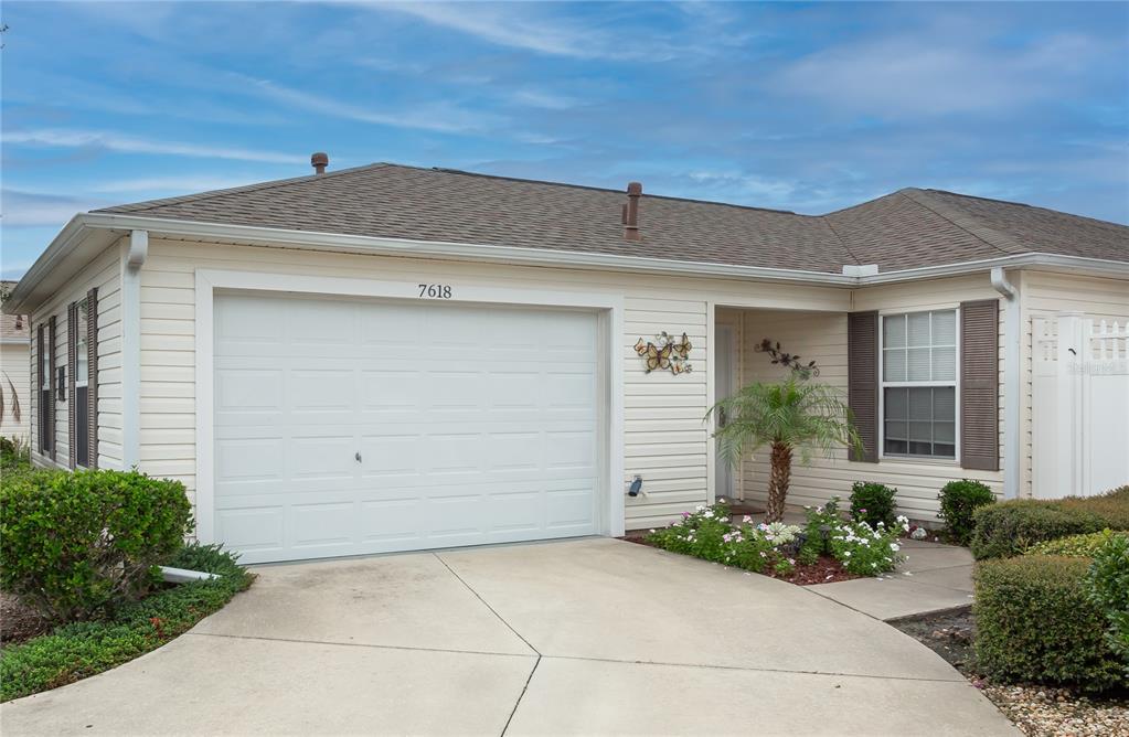 A covered front porch features a white door, a wicker chair, and decorative wall hangings — 7618 SE 171ST SUN VALLEY PLACE, THE VILLAGES, FL