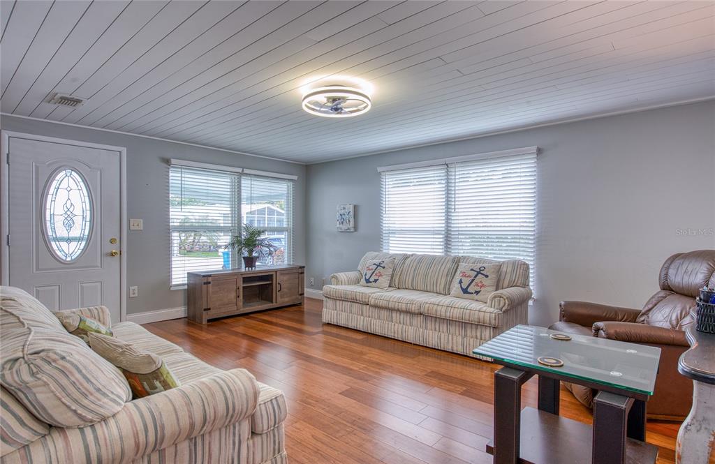 A living room features wood flooring, a white plank ceiling, two striped sofas, and blinds — 407 DOLPHIN DRIVE S, OLDSMAR, FL