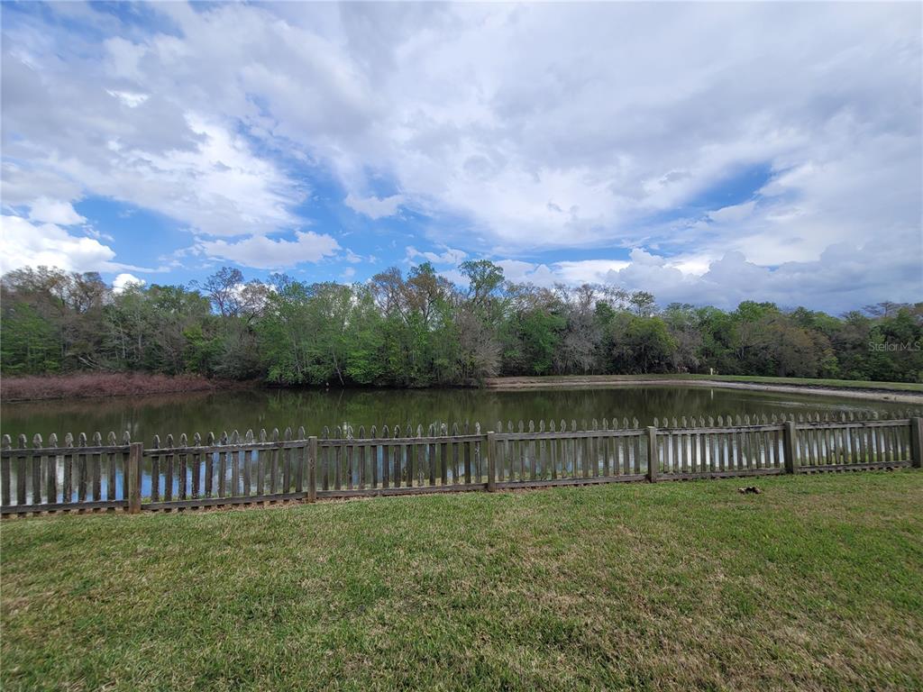 A wooden fence borders a pond with a treeline in the background under a cloudy sky — 27640 BREAKERS DRIVE, WESLEY CHAPEL, FL