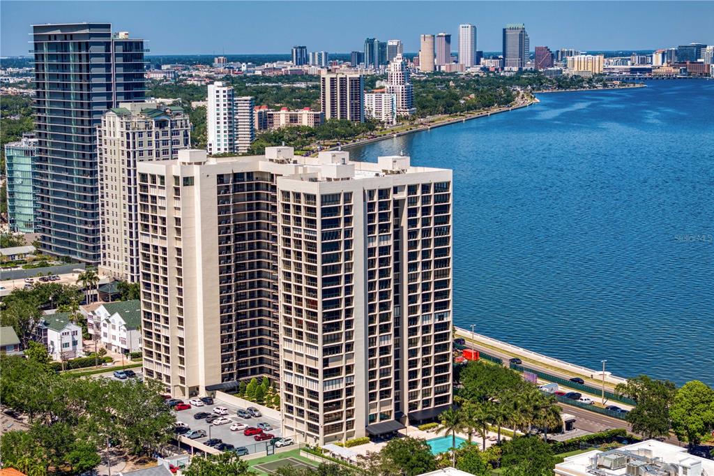 An aerial view shows a tall, beige apartment building situated along a waterfront with city skyscrapers — 3301 BAYSHORE BOULEVARD UNIT 509A, TAMPA, FL