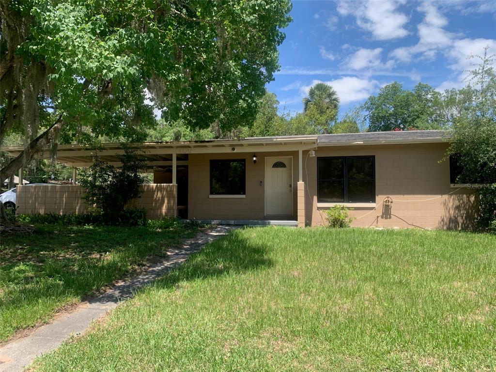 A single-story tan house with a carport and a front lawn under a large tree — 1406 PEMBROOK DRIVE, LEESBURG, FL