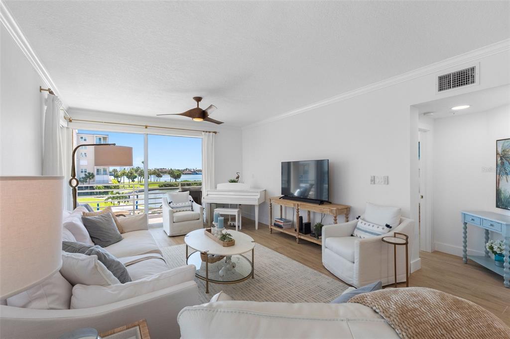 A bright living room features a white sofa, armchairs, a piano, and a waterfront balcony view — 7912 SAILBOAT KEY BOULEVARD S UNIT 201, SOUTH PASADENA, FL
