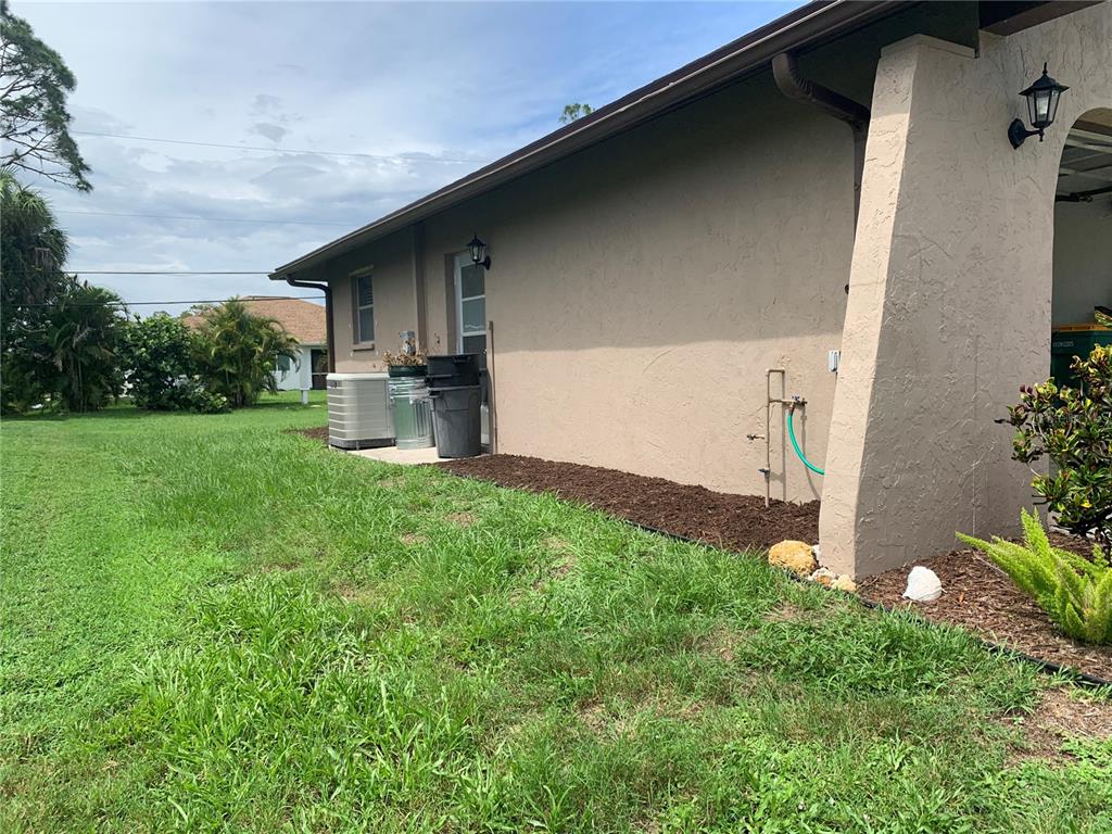 The exterior side of a house features a stucco wall, a small garden bed, and an HVAC unit — 18094 ACKERMAN AVENUE, PORT CHARLOTTE, FL