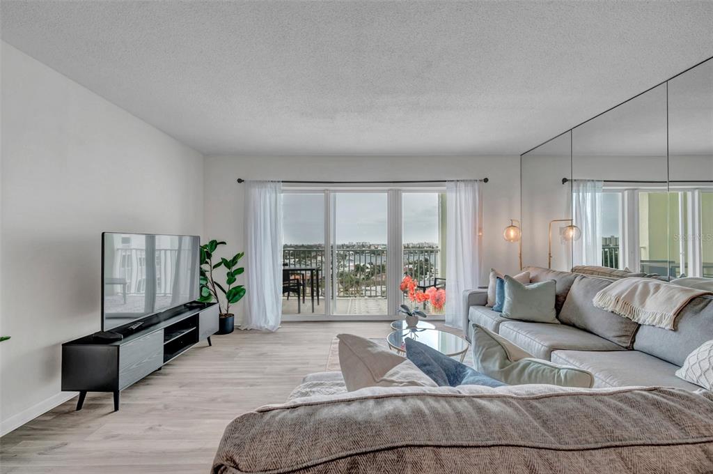 A living room features light wood flooring, a mirrored wall, and a balcony with water views — 400 64TH AVENUE UNIT 907 W, ST PETE BEACH, FL