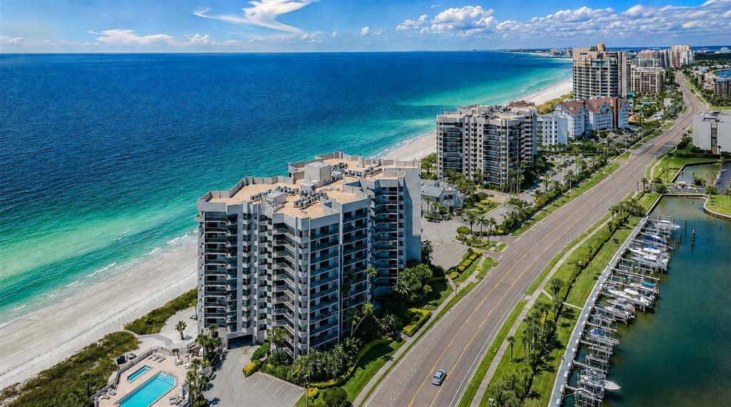 An aerial view shows a beachfront high-rise condominium building next to a road and marina — 1600 GULF BOULEVARD UNIT 1013, CLEARWATER, FL