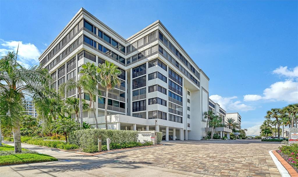 A multi-story residential building with screened balconies stands behind a paved driveway and palm trees — 1100 BENJAMIN FRANKLIN DRIVE UNIT 204, SARASOTA, FL