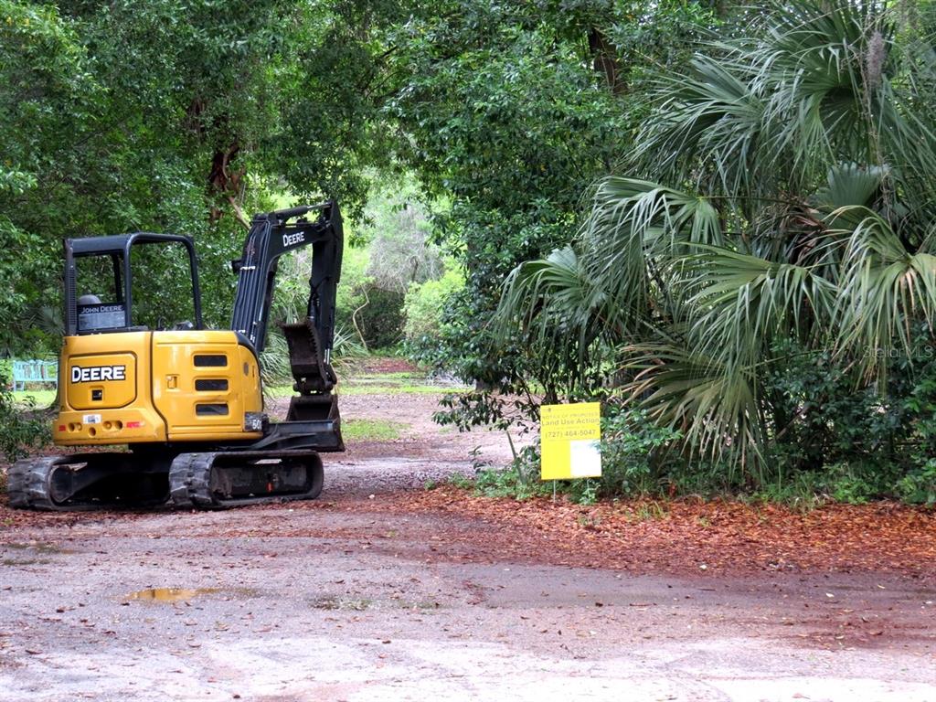 A yellow John Deere excavator sits on a dirt lot next to a land use sign — ROBERTS ROAD, PALM HARBOR, FL