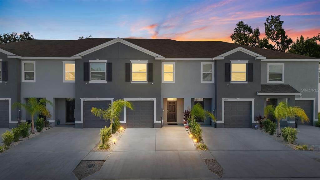 A multi-unit townhouse building features gray siding, attached garages, and small palm trees at dusk — 5052 CAPTAIN DAVIS DRIVE, WIMAUMA, FL