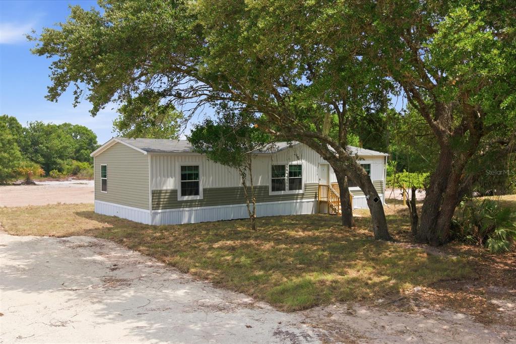 A single-story manufactured home with two-tone siding sits on a sandy lot under large trees — 20213 NW 258TH STREET, OKEECHOBEE, FL