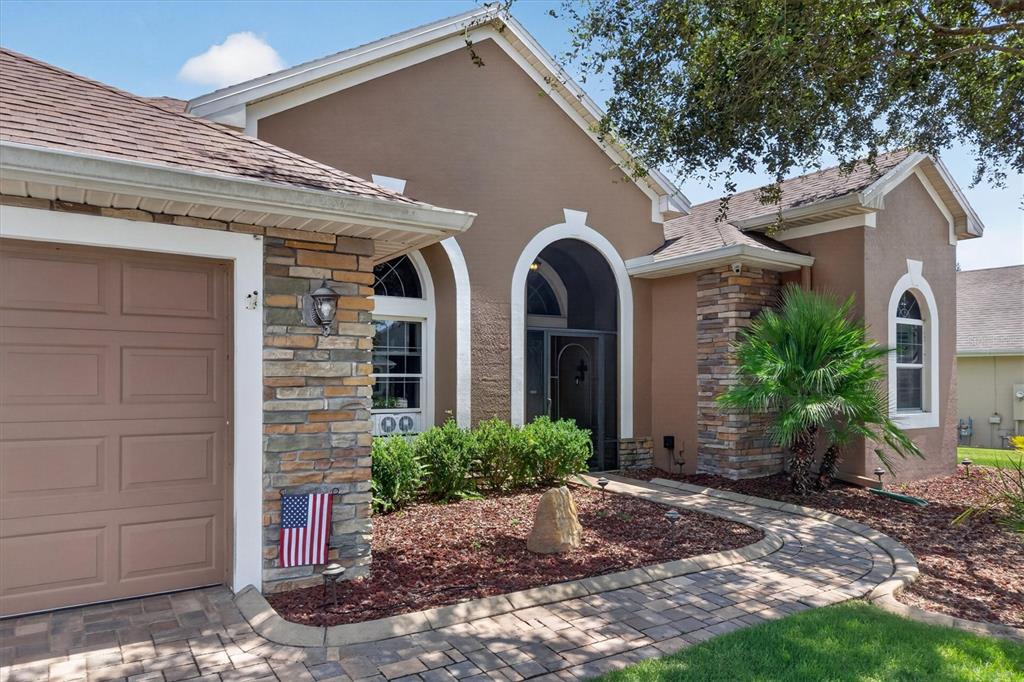 A tan house with stone accents, a paved walkway, and a garage under a blue sky — 8920 BRIDGEPORT BAY CIRCLE, MOUNT DORA, FL