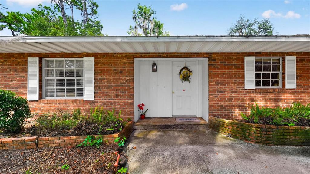 A brick house exterior features a white front door, two windows with shutters, and landscaping — 16529 ORANGE AVENUE, UMATILLA, FL