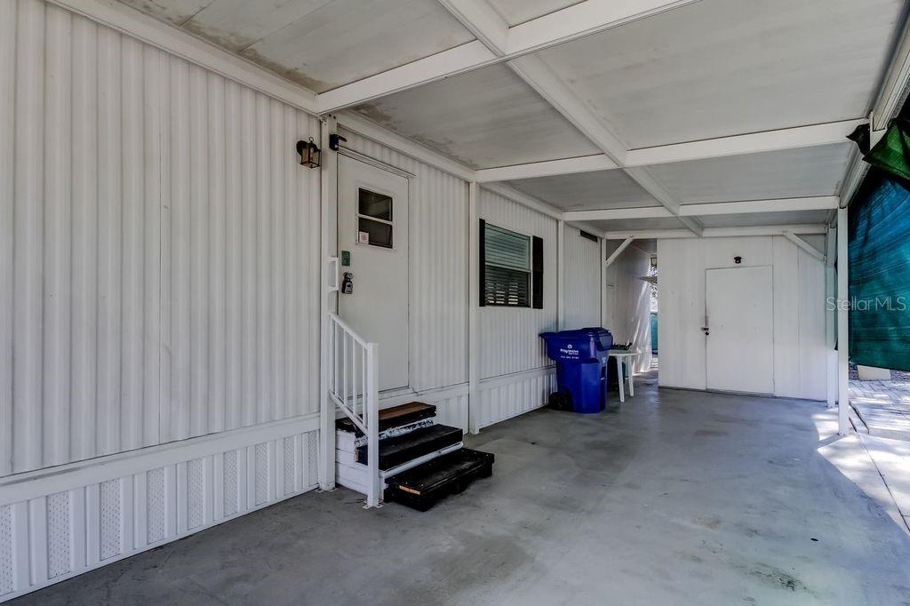A covered carport area features a white door with steps and a blue trash bin — 3430 TIKI DRIVE, HOLIDAY, FL