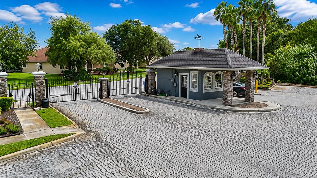 A gated entrance features a stone-accented guardhouse, paved driveway, and lush landscaping under a blue sky — 1637 ABYSS DRIVE, ODESSA, FL