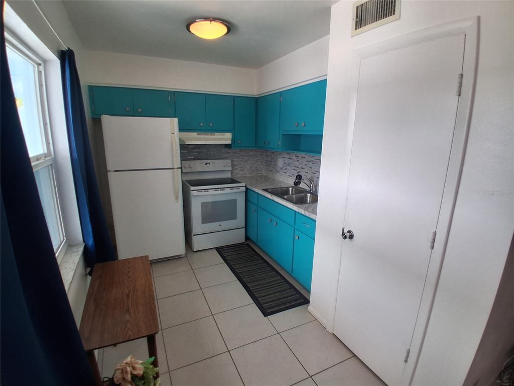 A kitchen features bright blue cabinets, white appliances, tiled flooring, and a window with curtains — 125 PORTREE DRIVE, DUNEDIN, FL
