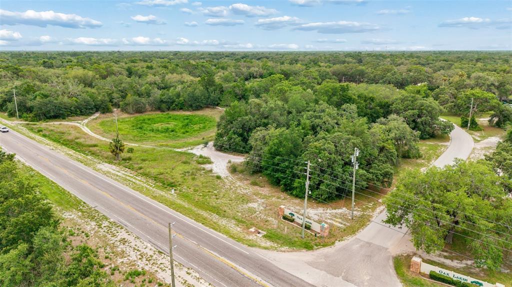 An aerial view shows a vacant, wooded lot with an entrance sign for Oak Lakes — COUNTY LINE ROAD, SPRING HILL, FL
