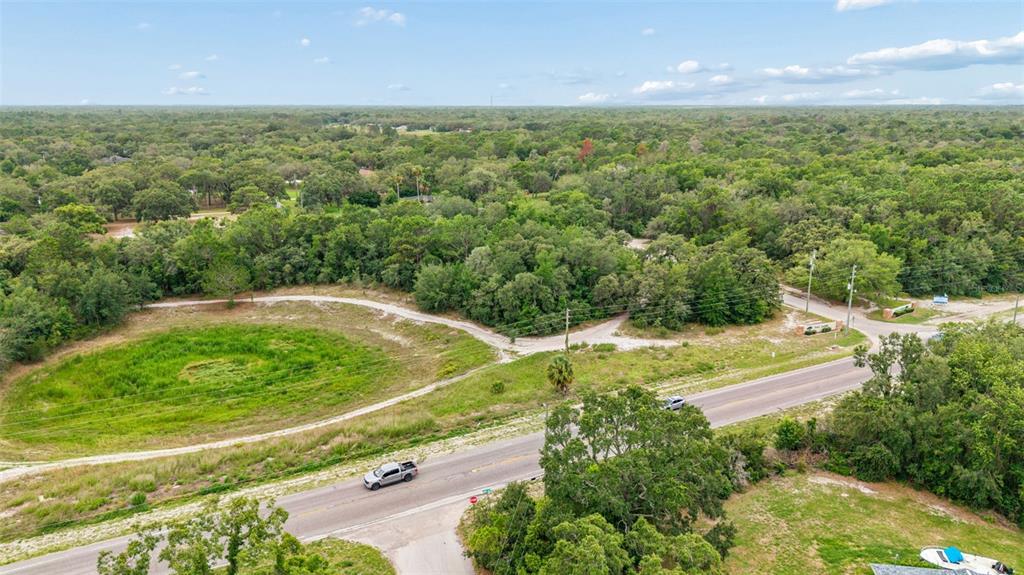 An aerial view shows a paved road running past a grassy lot and dense trees — COUNTY LINE ROAD, SPRING HILL, FL