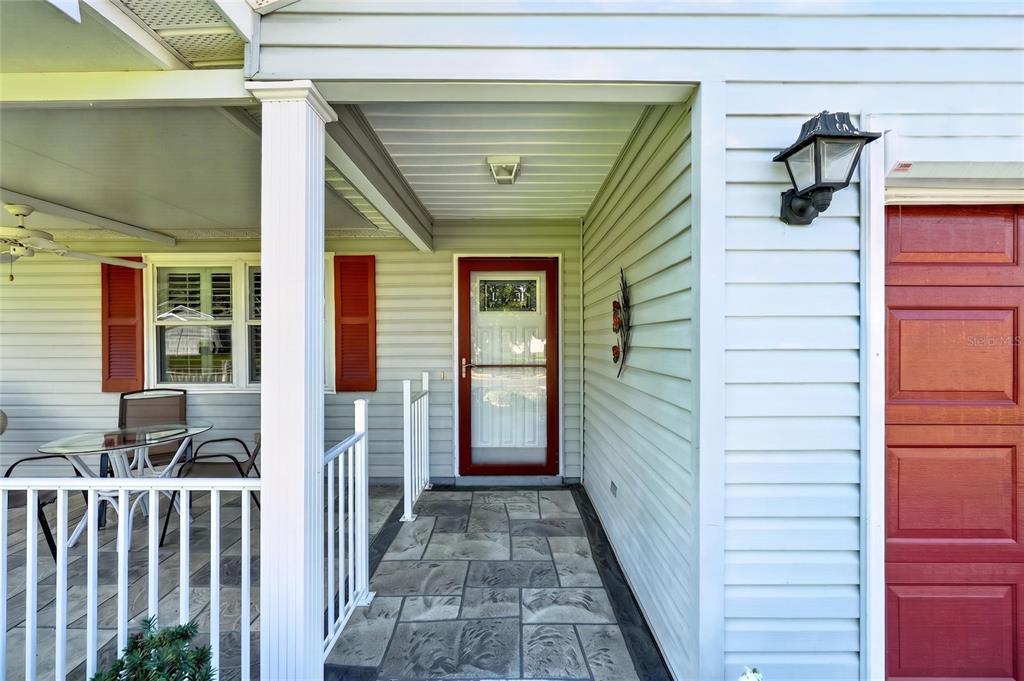 A covered front porch with a stone-patterned floor leads to a red-framed glass storm door — 1218 SANTOS PLACE, THE VILLAGES, FL