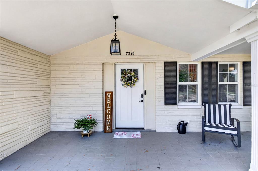 A covered front porch features a white door, a welcome sign, and a rocking chair — 7235 ASTOR DRIVE, NEW PORT RICHEY, FL