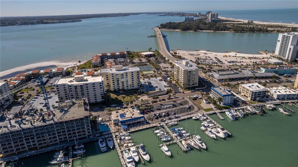 An aerial view shows a coastal marina with boats, multi-story buildings, and a long bridge — 830 S GULFVIEW BOULEVARD UNIT 501, CLEARWATER BEACH, FL