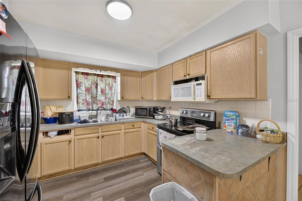 A kitchen features light wood cabinets, laminate countertops, stainless steel appliances, and a floral window curtain — 202 9TH STREET NE, RUSKIN, FL