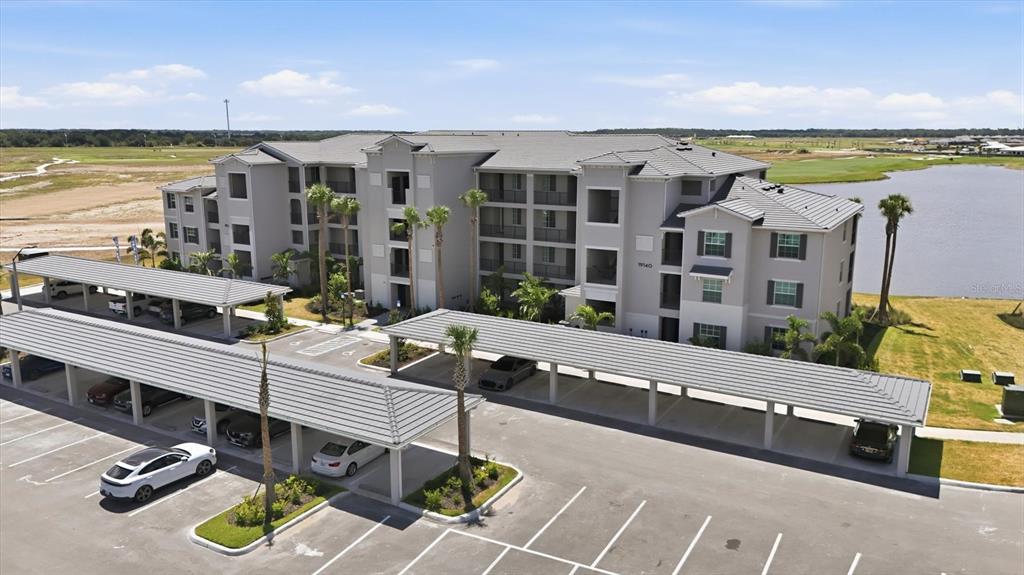 An aerial view shows a multi-story apartment building with covered parking structures and a lake — 19140 SCALLOP LOOP UNIT 108, LAKEWOOD RANCH, FL