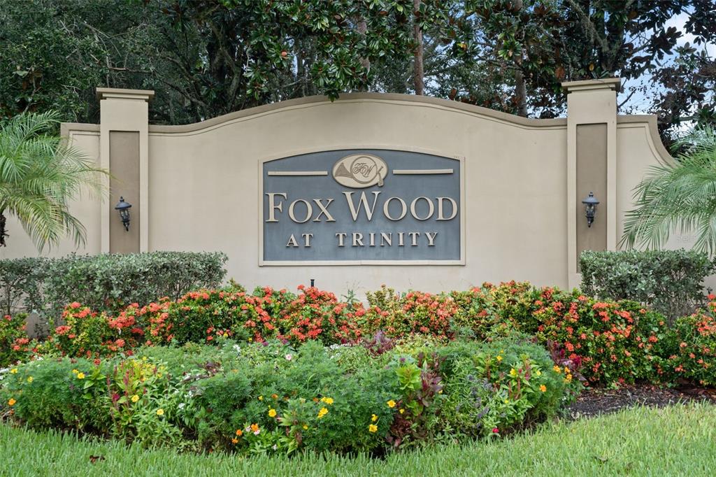 A beige monument sign for Fox Wood at Trinity sits behind a colorful garden bed — 2313 EDELWEISS LOOP, TRINITY, FL