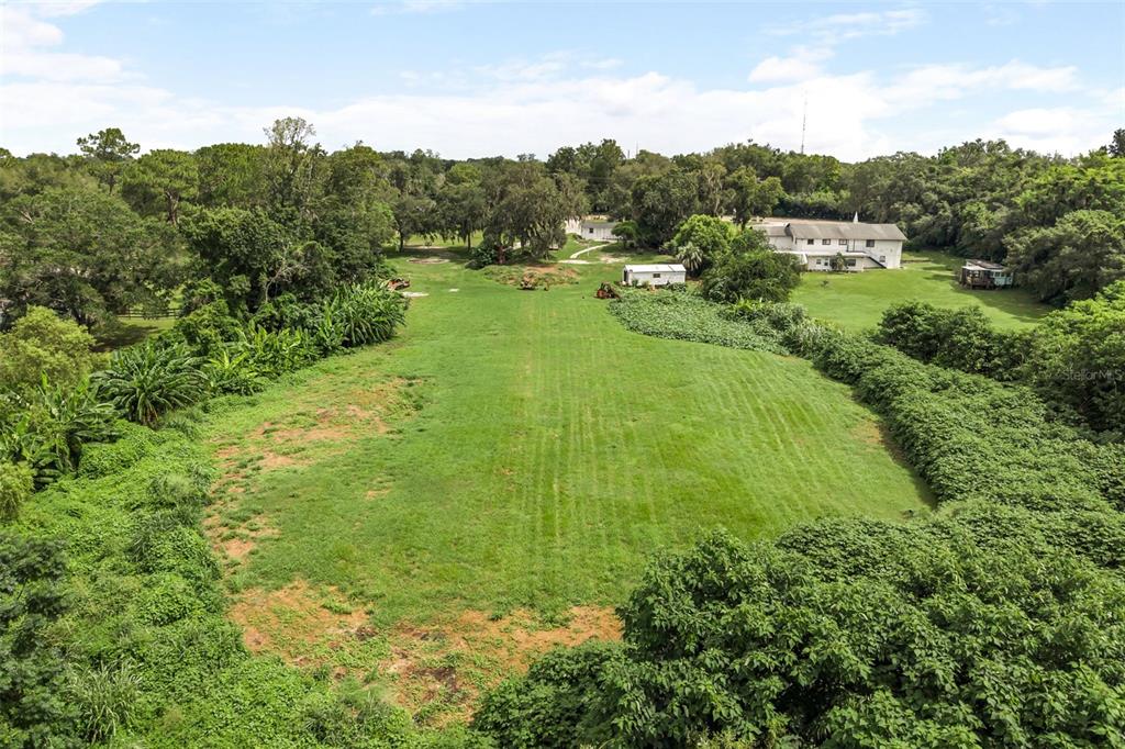 An aerial view shows a large, mowed grassy field surrounded by dense trees and several buildings — 250 LONGWOOD HILLS ROAD, LONGWOOD, FL