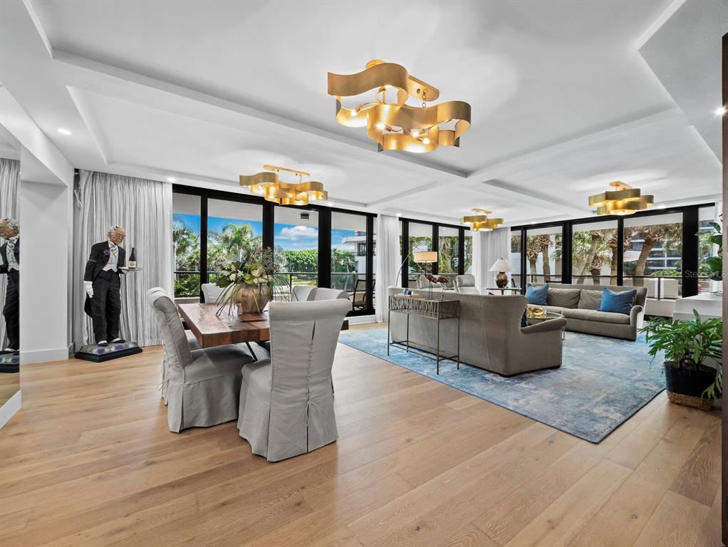 An open-concept living and dining area features wood floors, gold ribbon chandeliers, and floor-to-ceiling windows — 565 SANCTUARY DRIVE UNIT B106, LONGBOAT KEY, FL