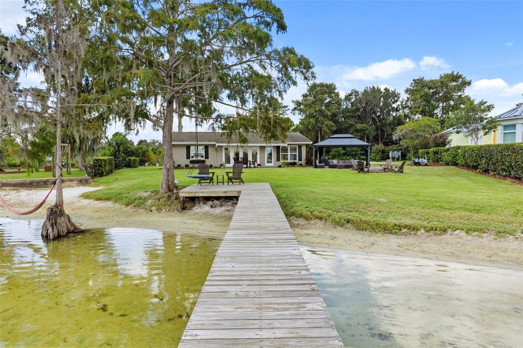 A wooden dock extends from a sandy shoreline toward a house with a backyard gazebo — 10430 LAKE COTTAGE LANE, ORLANDO, FL