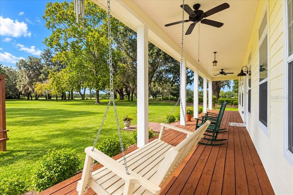 A covered wooden porch features a white swing, green rocking chairs, and a ceiling fan — 2451 NE 120TH STREET, CHIEFLAND, FL