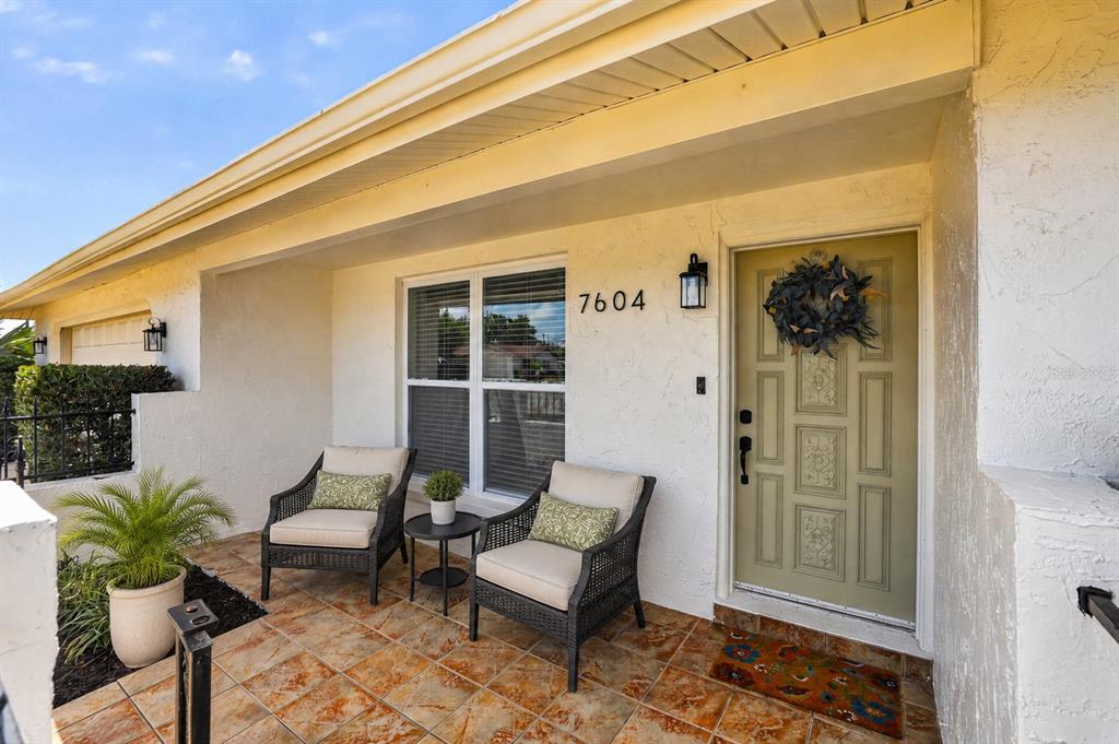 A covered front porch features tiled flooring, two wicker chairs, and a light green door — 7604 JASMINE BOULEVARD, PORT RICHEY, FL