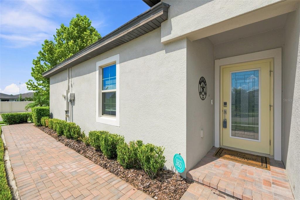 A light-colored house exterior features a yellow front door, a brick walkway, and green shrubs — 10444 WELDON CORK WAY, SAN ANTONIO, FL