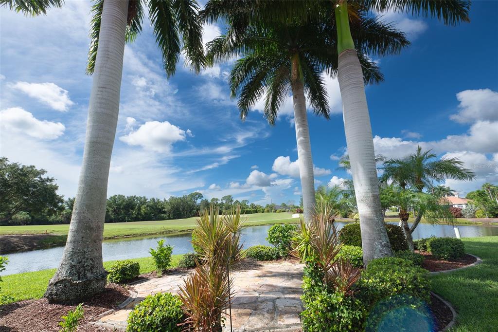 A stone patio sits beside a pond with tall palm trees under a blue sky — 11409 SW COURTNEY DRIVE, LAKE SUZY, FL